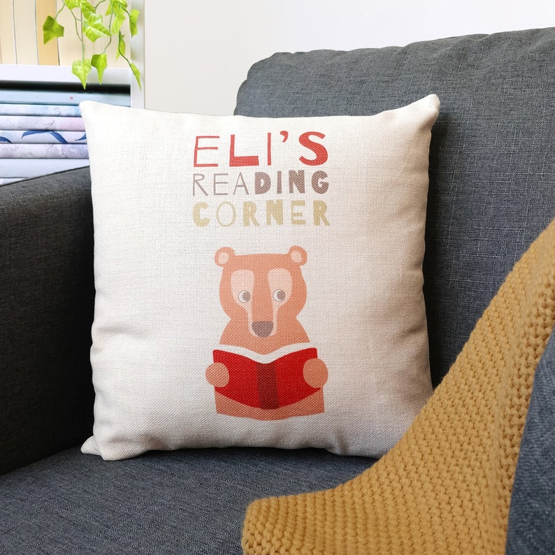 May include: A white throw pillow with the text "ELI'S READING CORNER" in brown, red, and beige lettering. A cartoon brown bear is holding a red book in front of it.
