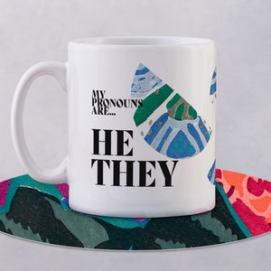 May include: White ceramic mug with a colourful abstract design and the text "My pronouns are... He They" printed in black.