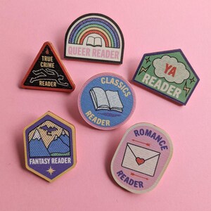Puede incluir: Un conjunto de parches bordados coloridos con textos como "Queer Reader", "True Crime Reader" y "Romance Reader". Los parches, de varias formas, presentan dise&ntilde;os como un arco&iacute;ris, un libro y un sobre con un coraz&oacute;n, sobre un fondo rosa.