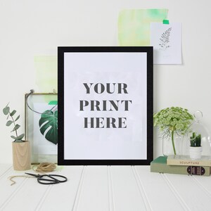 May include: A black picture frame with a white mat and the text "YOUR PRINT HERE" on a white background. The frame is sitting on a white table with a few plants and books.