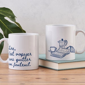 May include: Two white ceramic mugs with blue text and illustrations. The mug on the left says "Lire, c'est voyager sans quitter son fauteuil." The mug on the right has a drawing of a coffee pot, a cup of coffee, a croissant, and a book.