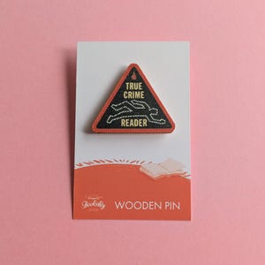 Puede incluir: Un pin de madera triangular con borde negro y las palabras "TRUE CRIME READER" en blanco. El pin presenta un contorno blanco de un cuerpo y est&aacute; colocado sobre una tarjeta blanca con una base naranja que dice "Bookishly WOODEN PIN".