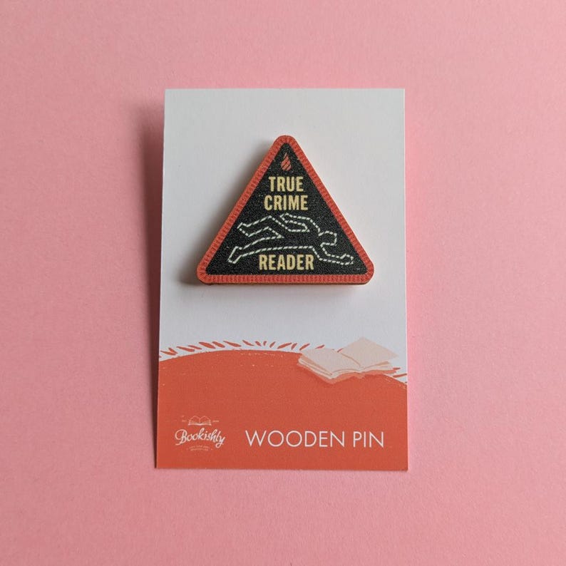 Puede incluir: Un pin de madera triangular con borde negro y las palabras "TRUE CRIME READER" en blanco. El pin presenta un contorno blanco de un cuerpo y est&aacute; colocado sobre una tarjeta blanca con una base naranja que dice "Bookishly WOODEN PIN".