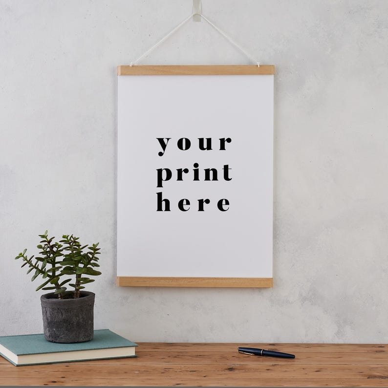 May include: A white framed poster with the text "your print here" hanging on a white wall. A small green plant is in a gray pot on a wooden table in front of the poster.