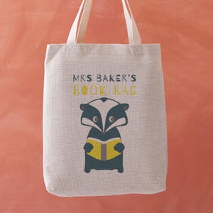 May include: A beige tote bag with cream-colored handles. The bag features the text "MRS BAKER'S BOOK BAG" in a combination of white and yellow. Below the text is a cartoon badger reading a book.