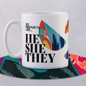 May include: White ceramic mug with a colorful abstract design and the text "My pronouns are... He She They" printed in black.