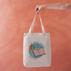 May include: Beige canvas tote bag with cream handles. The bag displays a circular graphic with a coffee cup, open book, and the text "SUNDAY READERS SOCIETY". The graphic uses teal, pink, and yellow.