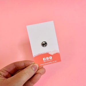 Puede incluir: Un pin plateado con cierre de mariposa, sujeto a una tarjeta blanca. La tarjeta tiene un dise&ntilde;o rosa y naranja en la parte inferior con iconos de redes sociales y la direcci&oacute;n del sitio web bookishly.co.uk.