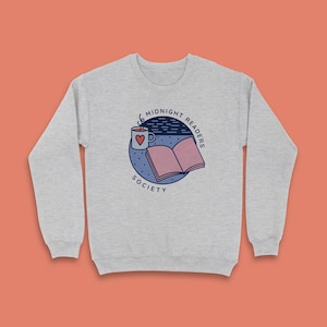 May include: A light grey sweatshirt with a circular graphic that reads "Midnight Readers Society." The design includes a mug with a heart, an open book, and a night sky. The sweatshirt has a crew neck.
