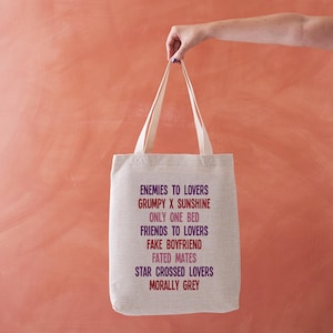 May include: A cream-coloured tote bag featuring phrases from romance novels, such as "ENEMIES TO LOVERS" and "GRUMPY X SUNSHINE," printed in red and purple. The bag has long, cream-coloured handles.