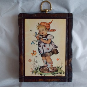 May include: A framed illustration of a young girl in a red and white dress holding a flower. The illustration is set against a background of flowers and butterflies. The frame is made of wood and has a gold-colored hanger.