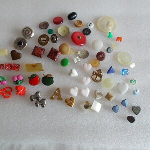 May include: A collection of assorted buttons in various colors, shapes, and sizes. The buttons are made of different materials, including plastic, metal, and fabric. Some of the buttons feature designs, such as hearts, anchors, and apples.