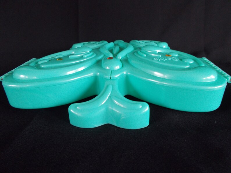 May include: A teal blue butterfly-shaped plastic trinket box with a raised design. The box is closed and has a small gold accent on the top.