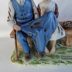 May include: A porcelain figurine of an older man and a young girl sitting on a bench. The man is wearing a blue vest and blue pants. The girl is wearing a white dress with a brown vest. The figurine is on a base with green grass and the words "Norman Rockwell" written on it.