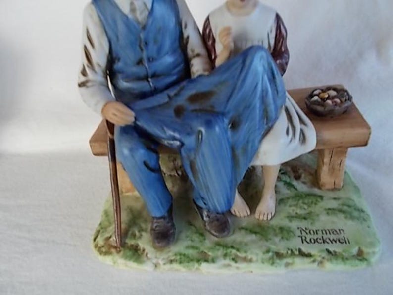 May include: A porcelain figurine of an older man and a young girl sitting on a bench. The man is wearing a blue vest and blue pants. The girl is wearing a white dress with a brown vest. The figurine is on a base with green grass and the words "Norman Rockwell" written on it.