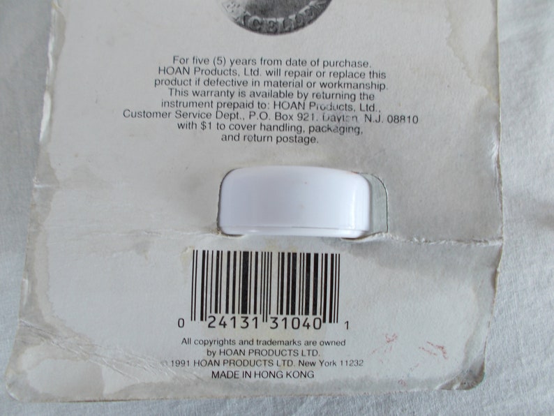 Op de afbeelding: Een wit plastic product met een barcode en tekst die luidt: "Voor vijf (5) jaar vanaf de aankoopdatum. HOAN Products. Ltd. zal dit product repareren of vervangen als het defect is in materiaal of afwerking. Deze garantie is beschikbaar door het instrument per vooruitbetaling terug te sturen naar: HOAN Products, Ltd.. Klantenservice, Postbus 921. Dayton N.J. 08810 met $ 1 voor afhandeling, verpakking en retourzending. 0 24131 31040 1 Alle auteursrechten en handelsmerken zijn eigendom van HOAN PRODUCTS LTD. 1991 HOAN PRODUCTS LTD. New York 11232 GEMAAKT IN HONGKONG"