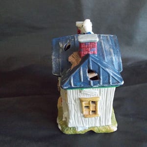 Może przedstawiać: Ceramiczna figurka domu z niebieskim dachem, czerwonym kominem z cegieł i białymi ścianami. Dom ma małe ż&oacute;łte okno i ozdobny komin z białą figurką na g&oacute;rze. Figurka jest pomalowana w stylu rustykalnym, postarzanym.