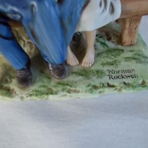 May include: A porcelain figurine of a couple sitting on a bench. The man is wearing blue pants and a brown jacket. The woman is wearing a white dress. The figurine is signed "Norman Rockwell".