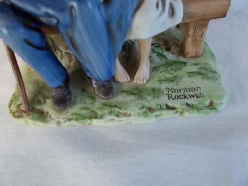 May include: A porcelain figurine of a couple sitting on a bench. The man is wearing blue pants and a brown jacket. The woman is wearing a white dress. The figurine is signed "Norman Rockwell".