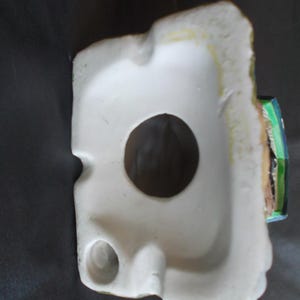 Może przedstawiać: Biała ceramiczna figurka z dużym okrągłym otworem i mniejszym otworem. Figurka ma szorstką, teksturowaną krawędź z odcieniami zielonej i ż&oacute;łtej farby. Mały fragment figurki jest pomalowany na zielono, niebiesko i brązowo.