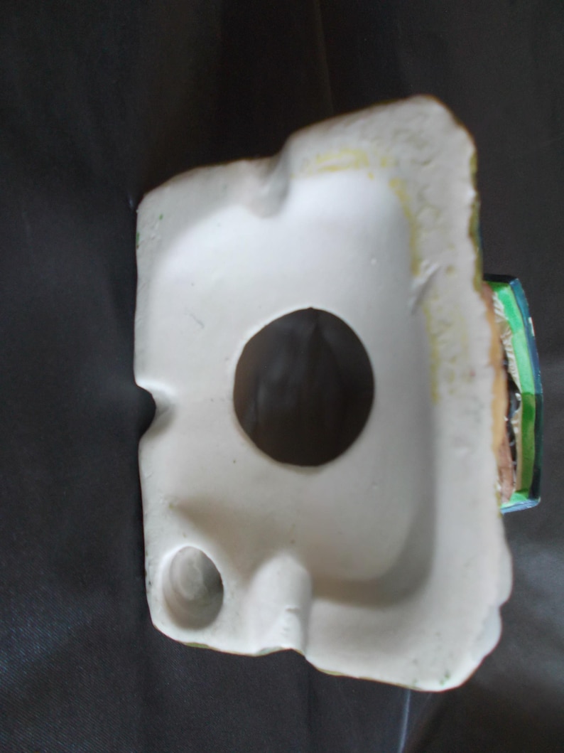 Może przedstawiać: Biała ceramiczna figurka z dużym okrągłym otworem i mniejszym otworem. Figurka ma szorstką, teksturowaną krawędź z odcieniami zielonej i ż&oacute;łtej farby. Mały fragment figurki jest pomalowany na zielono, niebiesko i brązowo.