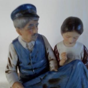 May include: A porcelain figurine set featuring a man wearing a blue cap and a blue vest, and a woman wearing a white dress with a red and white patterned bodice. The man is holding the woman's hand.