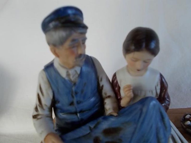 May include: A porcelain figurine set featuring a man wearing a blue cap and a blue vest, and a woman wearing a white dress with a red and white patterned bodice. The man is holding the woman's hand.