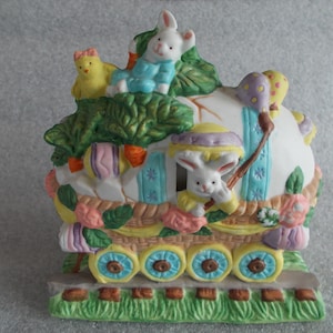 May include: A ceramic Easter egg train with a bunny, chick, and flowers. The train is decorated with pastel colors and has a white egg-shaped car.