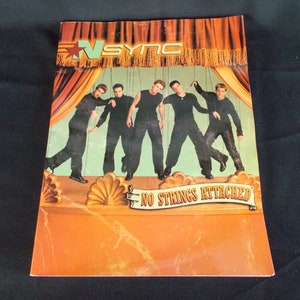 May include: A music album cover featuring a boy band, *NSYNC, with the title "No Strings Attached". The band members are dressed in black and are posed as puppets on a stage.