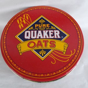 Op de afbeelding: Een ronde blik van rood metaal met een gouden en blauw Quaker Oats logo. Het logo heeft de vorm van een ruit met de woorden "Pure Quaker Oats" erin. De blik heeft een gouden rand met een herhalend patroon van kleine bloemen.