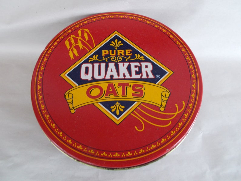 Op de afbeelding: Een ronde blik van rood metaal met een gouden en blauw Quaker Oats logo. Het logo heeft de vorm van een ruit met de woorden "Pure Quaker Oats" erin. De blik heeft een gouden rand met een herhalend patroon van kleine bloemen.