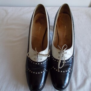 May include: A pair of two-tone, black and white oxford-style heels. The shoes have a pointed toe, a lace-up closure, and small decorative perforations. The heels are approximately 3 inches high.