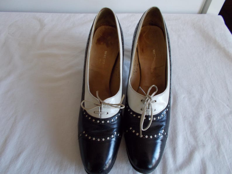May include: A pair of two-tone, black and white oxford-style heels. The shoes have a pointed toe, a lace-up closure, and small decorative perforations. The heels are approximately 3 inches high.