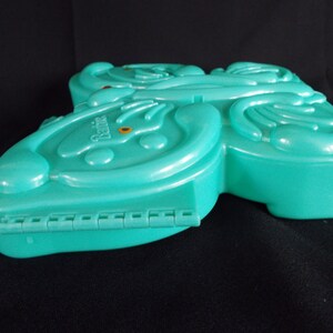 May include: A teal blue plastic butterfly-shaped case with the word "Barbie" printed on the front. The case has a hinge on the bottom and is closed.