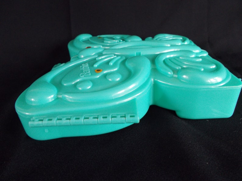 May include: A teal blue plastic butterfly-shaped case with the word "Barbie" printed on the front. The case has a hinge on the bottom and is closed.