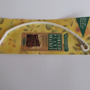 May include: A white, curved forged plant bracket on a yellow package with floral designs. The bracket is designed for outdoor use and includes mounting screws. Text on the package reads "Forged Plant Bracket" and "Weather-Resistant for Outdoor Use."