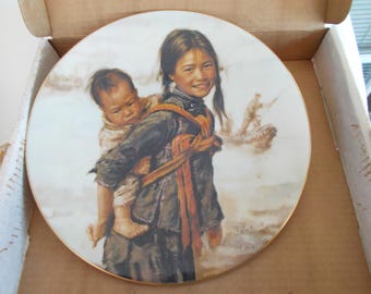 Op de afbeelding: Decoratieve plaat met een schilderij van een jonge persoon die een baby draagt. De plaat heeft een gouden rand en toont een persoon in een boot op de achtergrond. De plaat zit in een kartonnen doos.