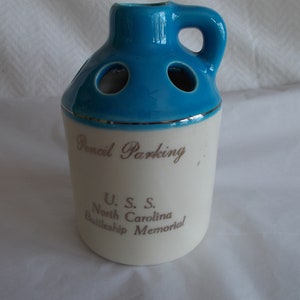 May include: A white ceramic pencil holder shaped like a jug with a blue top and a silver trim. It says "Pencil Parking" and "U.S.S. North Carolina Battleship Memorial" on the front.