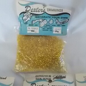 May include: A package of Dexter's Trimmings, featuring small, golden-yellow beads. The packaging includes the text "DECORATE YOUR OWN" and the number "16-53062". The beads are suitable for decorating dresses, hats, and other items.