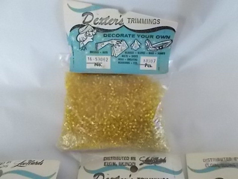 May include: A package of Dexter's Trimmings, featuring small, golden-yellow beads. The packaging includes the text "DECORATE YOUR OWN" and the number "16-53062". The beads are suitable for decorating dresses, hats, and other items.