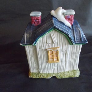 Może przedstawiać: Ceramiczny miniaturowy domek z białym zewnętrzem, niebieskim dachem i czerwonymi ceglanymi kominami. Dom ma małe ż&oacute;łte okno i zieloną obw&oacute;dkę. Ozdobny akcent śnieżny znajduje się na dachu.