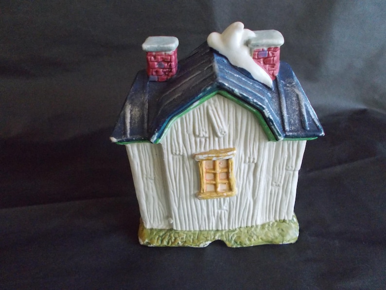 Może przedstawiać: Ceramiczny miniaturowy domek z białym zewnętrzem, niebieskim dachem i czerwonymi ceglanymi kominami. Dom ma małe ż&oacute;łte okno i zieloną obw&oacute;dkę. Ozdobny akcent śnieżny znajduje się na dachu.