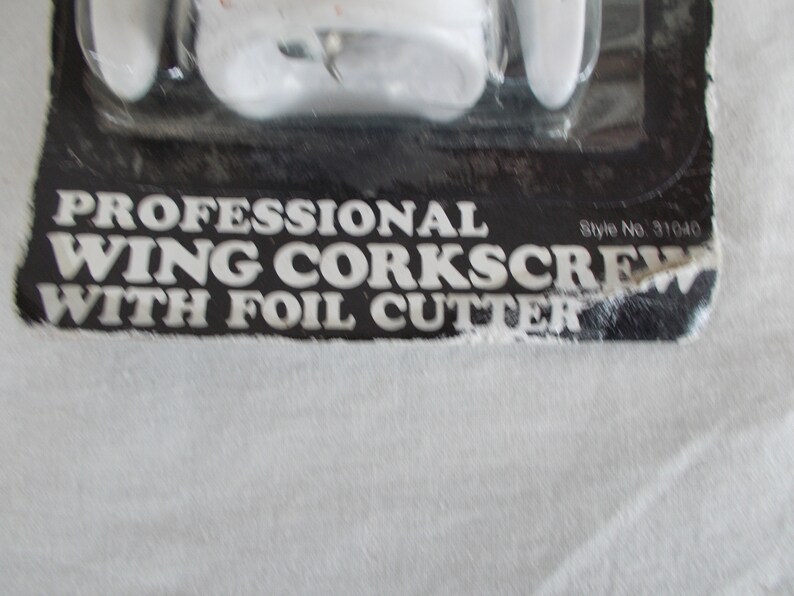 Op de afbeelding: Een professionele vleugelkurkentrekker van wit plastic met een folie-snijder. De verpakking is zwart met witte tekst die "Professional Wing Corkscrew with Foil Cutter" en "Style No. 31040" luidt.