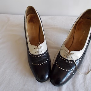 May include: A pair of vintage black and white leather oxford-style shoes. The shoes have a pointed toe, white laces, and decorative perforations along the seams. The heels are low and the shoes appear to be in good condition.