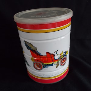 May include: A vintage white metal canister with a red and yellow stripe design. The canister features a colorful illustration of a red Cadillac Surrey Type touring car. The lid is white plastic with the text "Cadillac Surrey Type" printed in black.