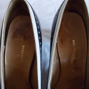 May include: Pair of black and white leather shoes. The shoes have a classic design with a white trim and small decorative holes. The interior of the shoes is a light brown color, with the words "SHOE BIZ" printed on the insole.