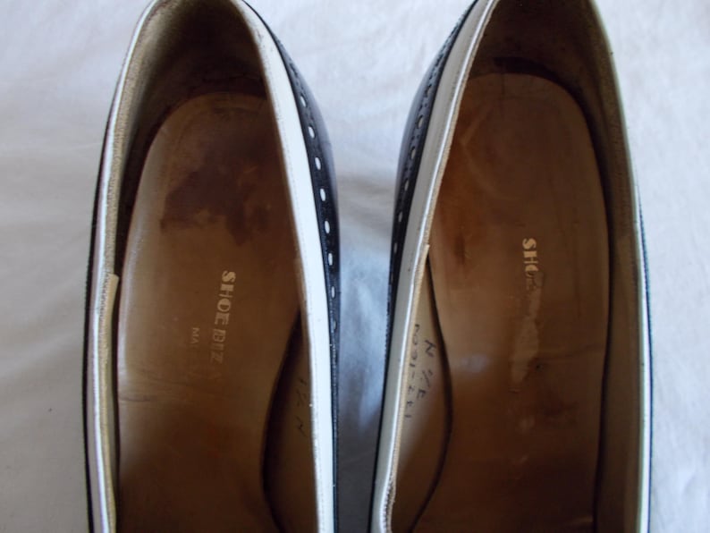 May include: Pair of black and white leather shoes. The shoes have a classic design with a white trim and small decorative holes. The interior of the shoes is a light brown color, with the words "SHOE BIZ" printed on the insole.