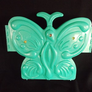 May include: A teal blue plastic butterfly-shaped jewelry box with gold accents. The box has the word "Barbie" printed on the front.