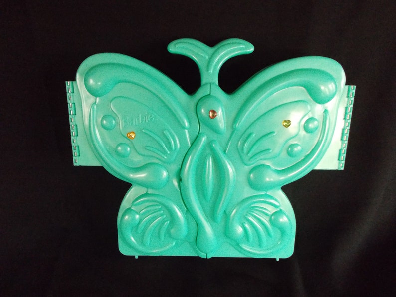 May include: A teal blue plastic butterfly-shaped jewelry box with gold accents. The box has the word "Barbie" printed on the front.