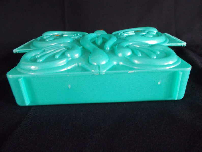May include: A teal plastic box with a decorative butterfly design on the lid. The box is closed and the lid is slightly ajar.